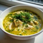 Close-up of a white bowl filled with bright yellow Mediterranean lemon chicken soup, rice, chicken pieces, and topped generously with fresh dill.