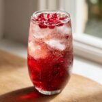 A tall glass filled with a vibrant red Pomegranate spritz cocktail, ice, bubbles, and topped with fresh pomegranate seeds.