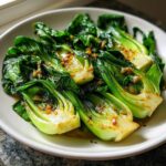 Close-up of vibrant green sautéed bok choy with garlic pieces glistening in sauce on a white plate.